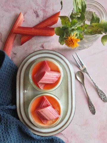 Panna cotta met rabarber in twee glazen schaaltjes die op een bord staan. De toetjes zijn gegarneerd met stukken gestoofde rabarber.