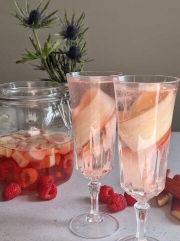 Twee glazen rabarber cocktail, versierd met linten rabarber. Er staat een rabarberdrank op de achtergrond.