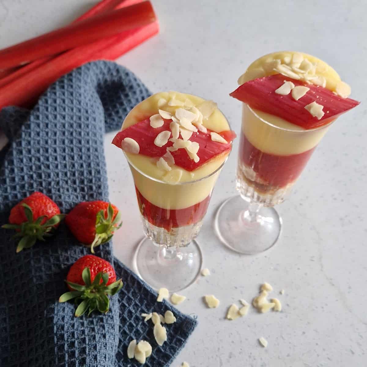 Bovenaanzicht van twee glazen gevuld met rabarbertrifle. Er liggen verse aardbeien en amandelschaafsel naast.