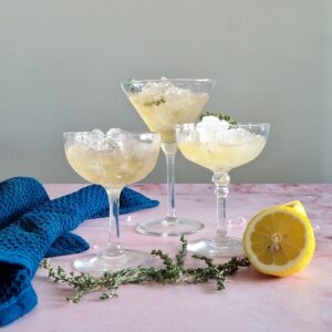 Lavendel cocktail in drie verschillende cocktailglazen, die gevuld zijn met veel crushed ice. Er liggen takjes tijm en een gehalveerde citroen voor.