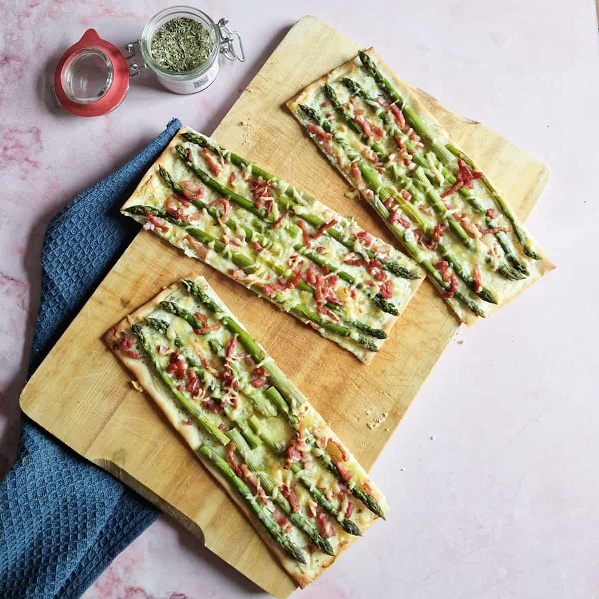 In drie stukken gesneden flammkuchen met asperges en spekjes, op een houten serveerplank.