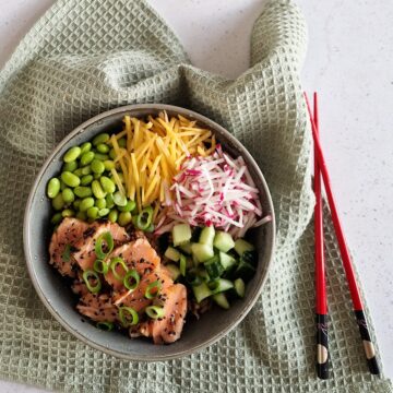Sushibowl met teriyaki zalm, edamame, wortel, radijs en komkommer. Er liggen eetstokjes naast de kom.