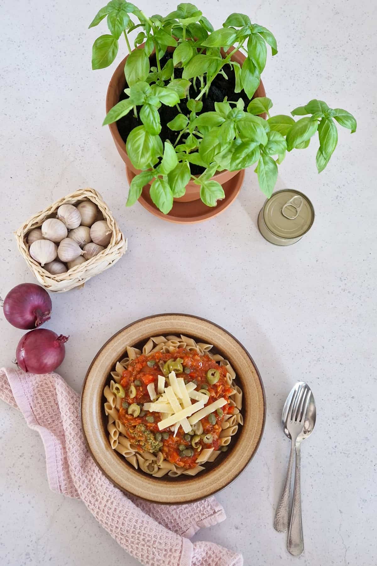 een bord pasta met bestek, een basilicumplant, teentjes knoflook en uien