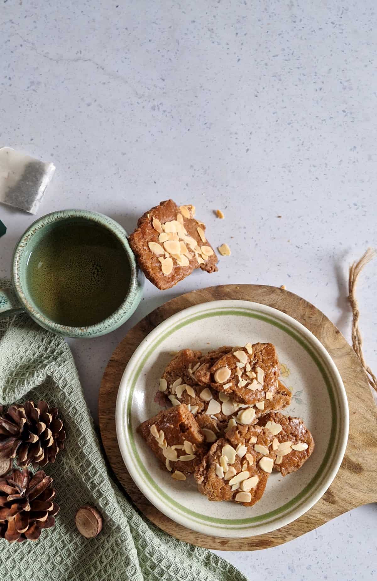 Dikke speculaasbrokken met amandel, op een gebaksbordje. Erbij een kopje koffie en wat dennenappels ter decoratie.