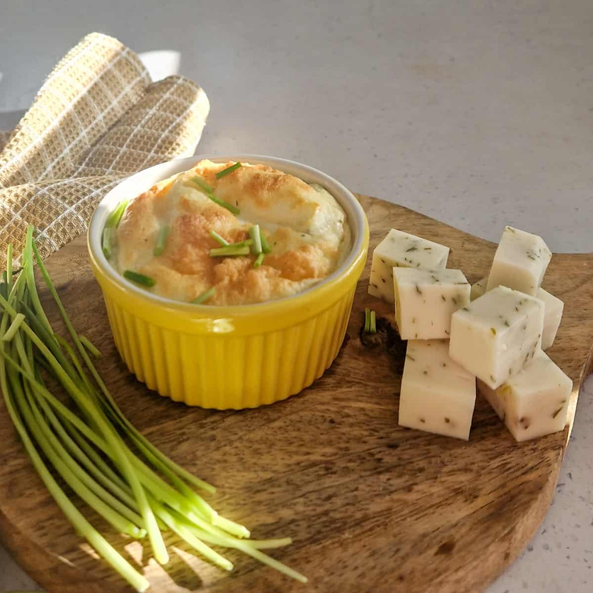 Een klein borrelplankje met een schaaltje met geitenkaassoufflé. Er omheen blokjes kruidige geitenkaas en sprieten bieslook.