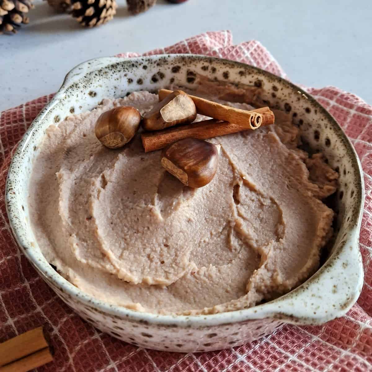 Een kom met gladde kastanjepuree. Er liggen twee kaneelstokjes en drie kastanjes op ter decoratie.