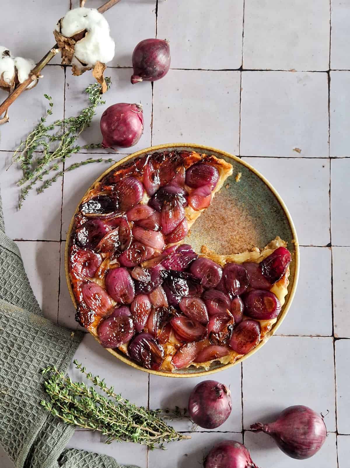 Een tarte tatin van rode uien op een bord, met een puntje eruit gesneden. Er omheen de ingrediënten van het recept: rode ui en tijm.