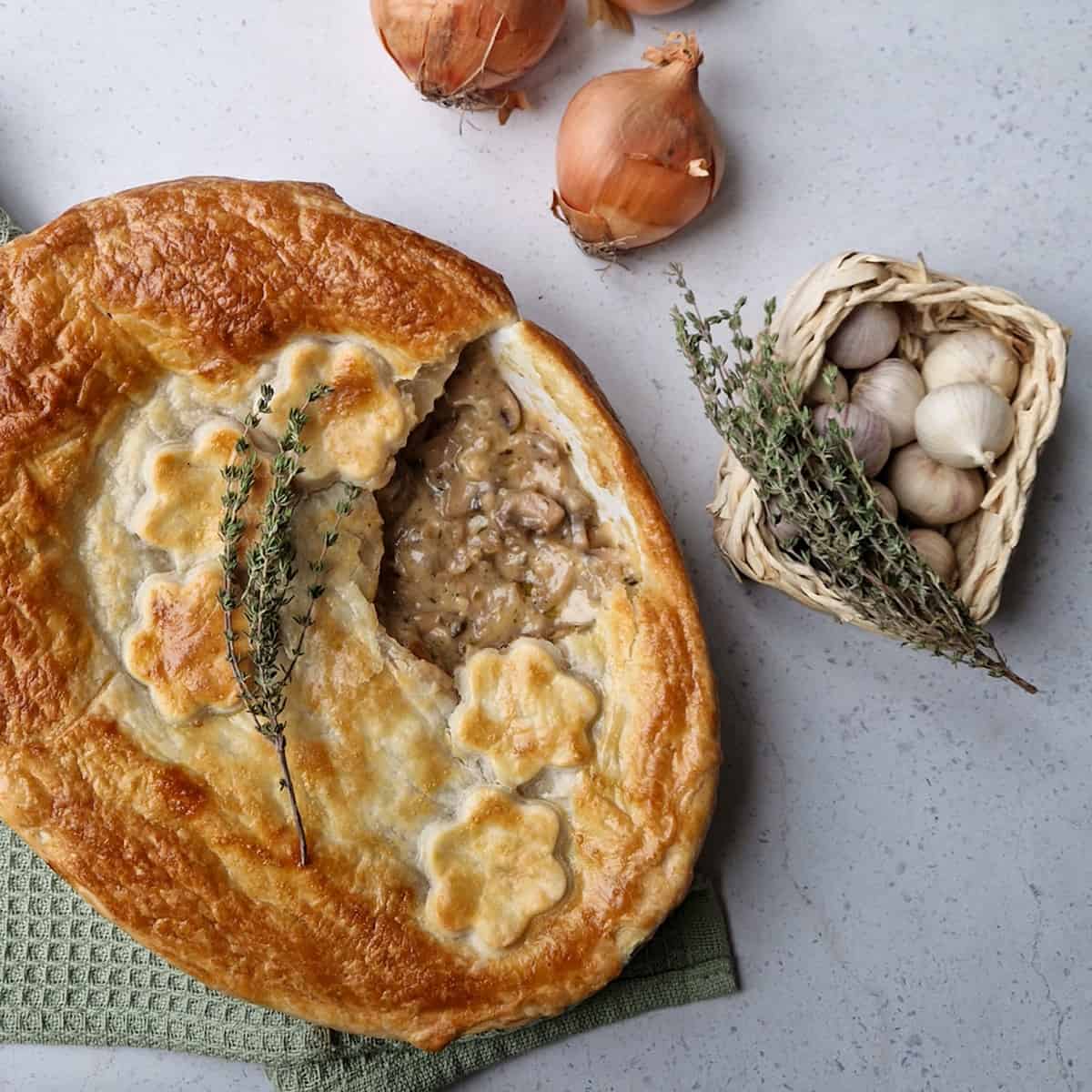 Een ovenschotel met een vegetarische pastei met paddenstoelen. Ter decoratie liggen er uien, knoflook en tijm bij.