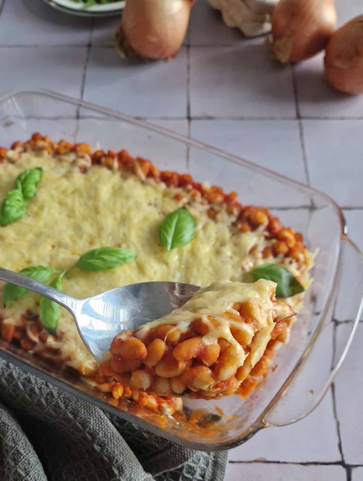 Bonenschotel van witte bonen met tomatensaus, een hand schept een lepel uit de bonenschotel, zodat je de inhoud goed kunt zien.