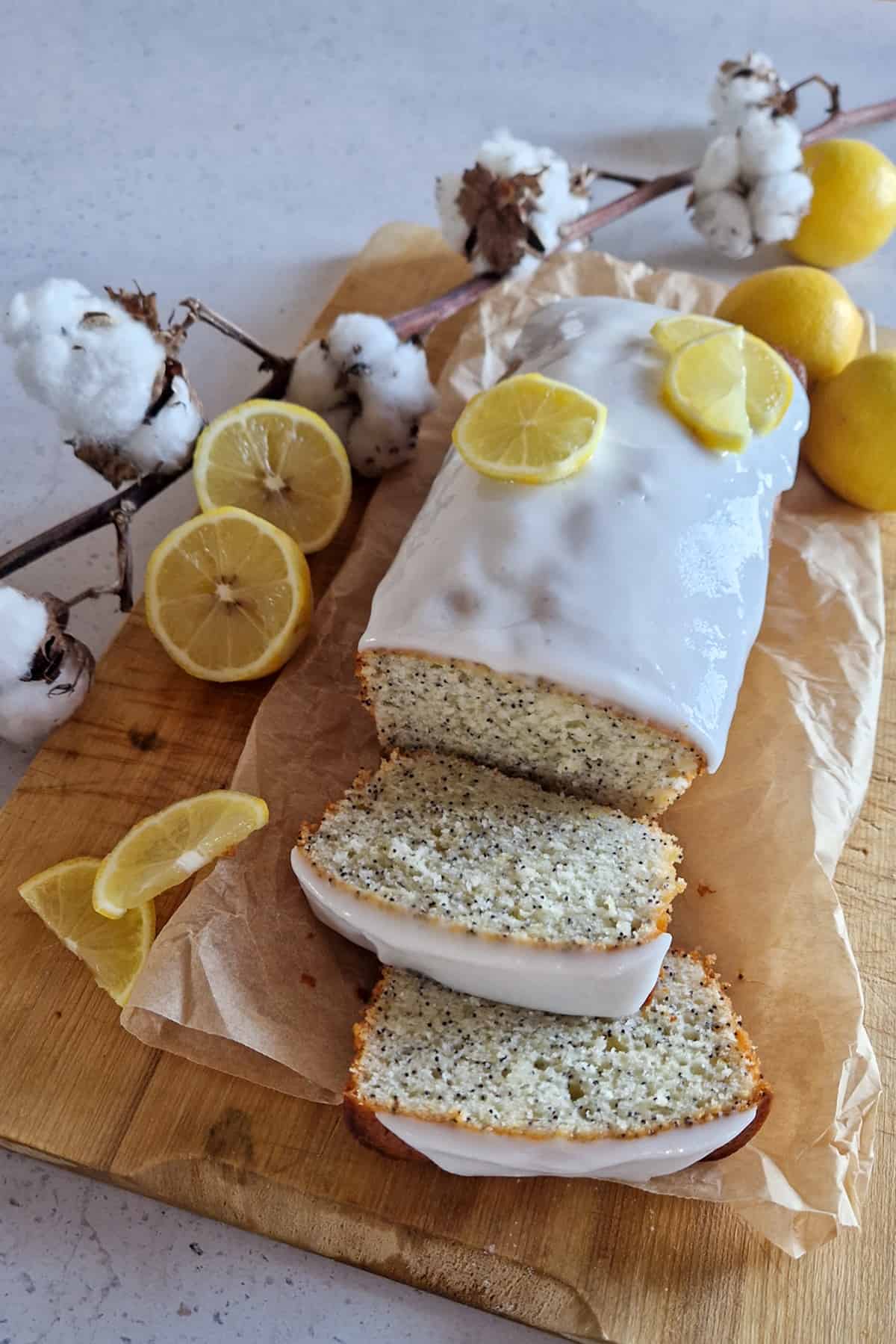 Een citroencake met maanzaad waar twee plakken vanaf zijn gesneden. De cake ligt op een houten plank op bakpapier en is gegarneerd met een witte frosting en schijfjes citroen.