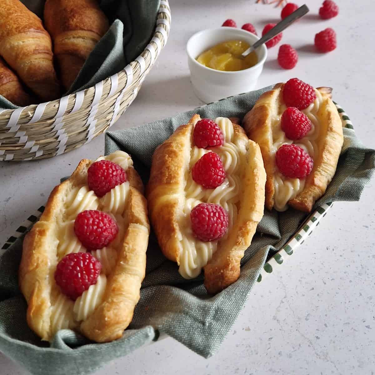 Drie afbakcroissants met citroenroom en frambozen. De broodjes liggen op een bord met een groene doek.