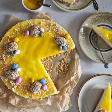 Paastaart met lemon curd en paaschocolade-decoraties. Uit de taart is een flinke punt gesneden, die in drie kleine puntjes op drie gebaksbordjes om de taart heenstaan.