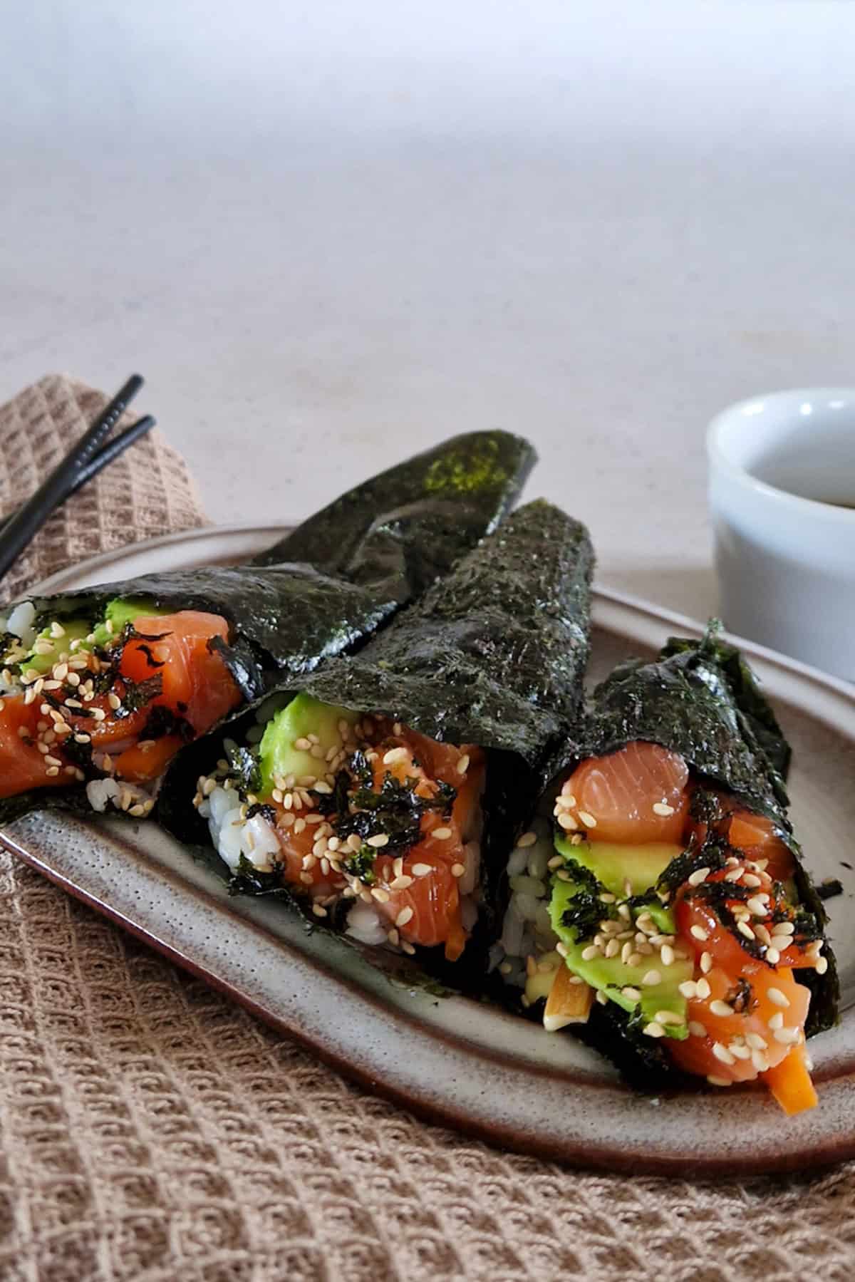 Drie handrolls met zalm op een bordje. Ernaast liggen eetstokjes en een bakje met sojasaus, om de handrolls in te dippen.