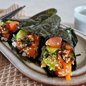 Temaki sushi met zalm, avocado en wortel. De handrolls liggen met drie naast elkaar op een bordje en zijn besprenkeld met nori en sesamzaadjes.