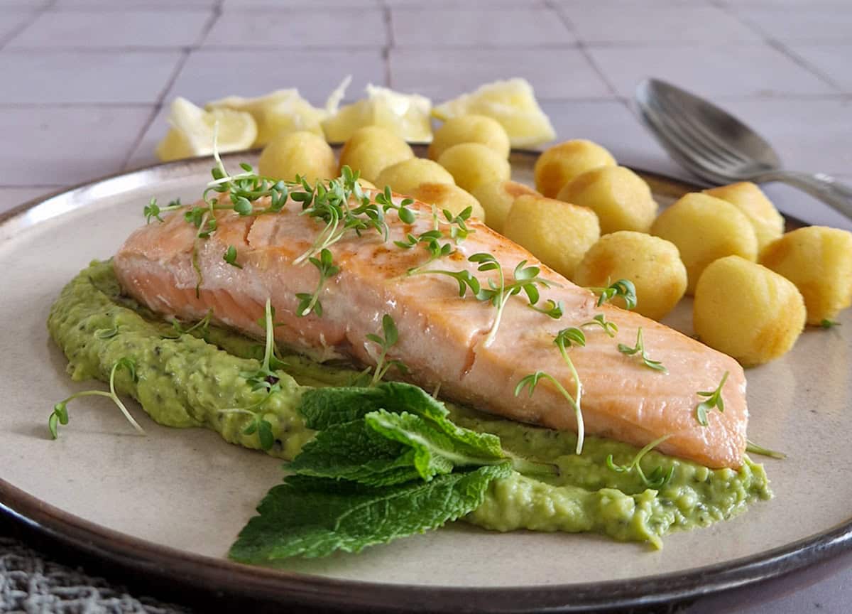 Een stukje mooi gebakken zalm op een bedje doperwtenpuree. De zalm wordt geserveerd met aardappels en citroen.