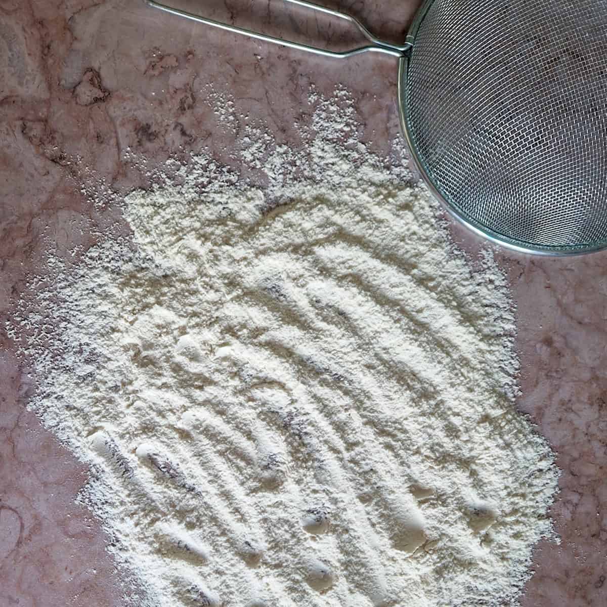 Een uitgewaaierde hoeveelheid zelfgemaakt zelfrijzend bakmeel met een zeef ernaast.