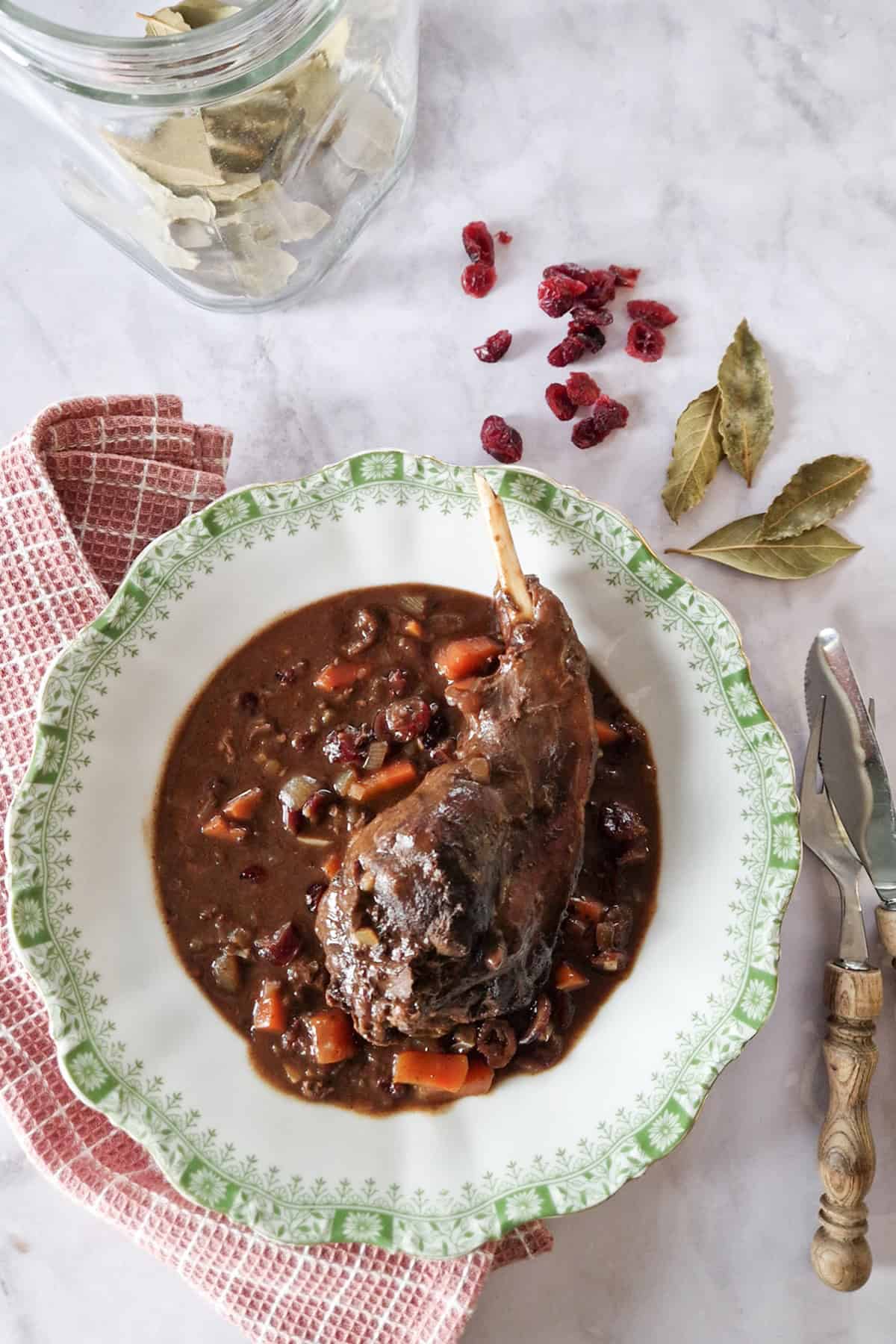 Een bord ouderwetse hazenpeper, omringd door bestek, cranberries en laurierblaadjes.