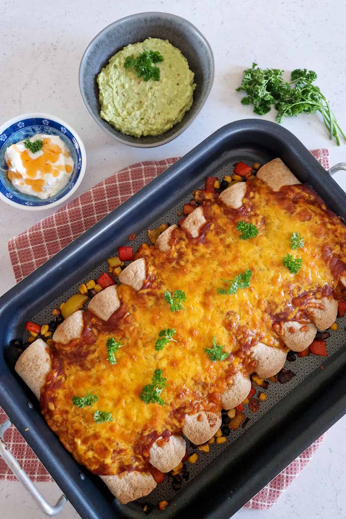 Enchilada recept met bonen en veel verse groenten. De enchiladas zijn overgoten met saus en geraspte cheddar, en worden geserveerd met guacemole en zure room in schaaltjes naast de ovenschotel.
