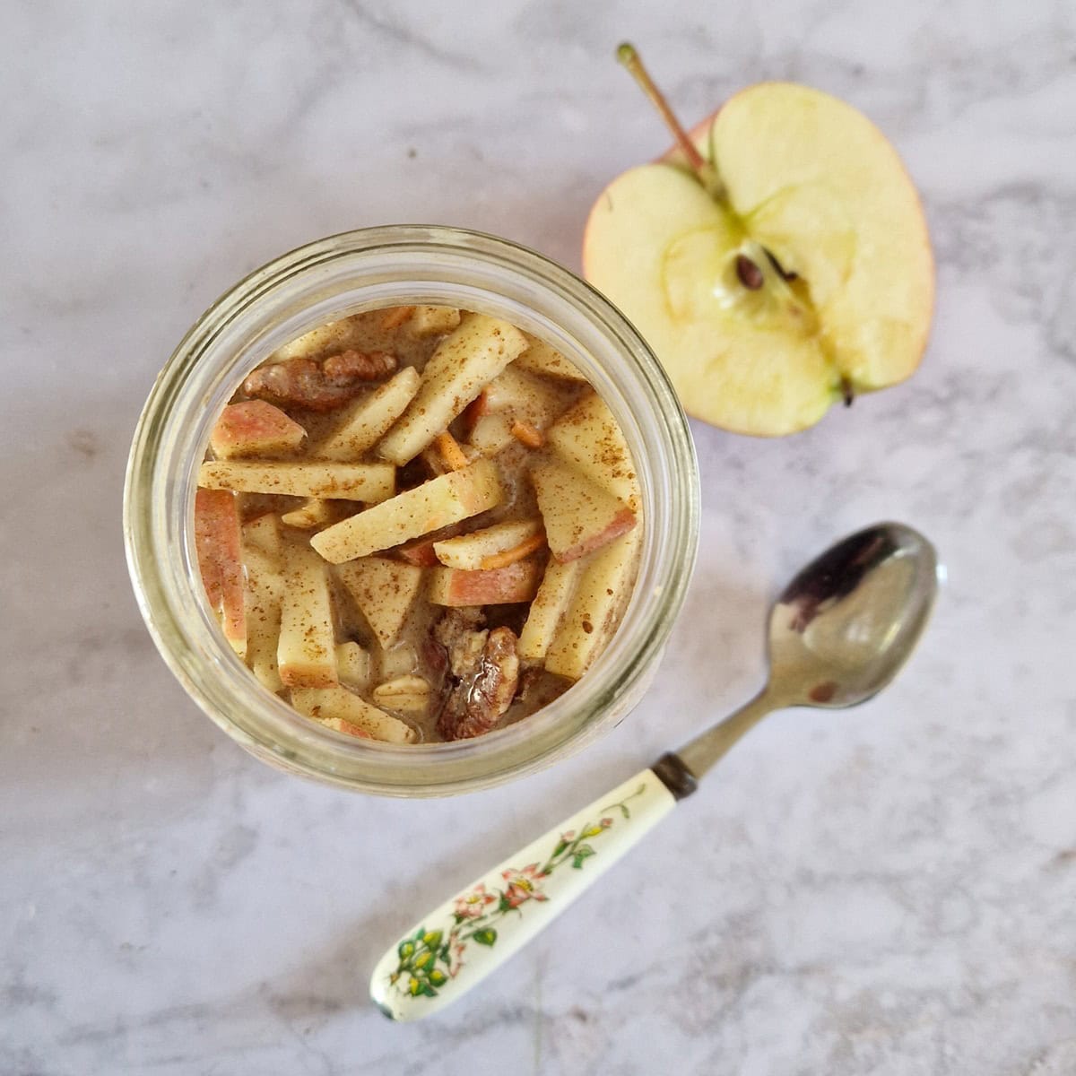 Bovenaanzicht van een glas met carrot cake overnight oats. Ernaast een lepel en een halve appel.
