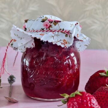 Zelfgemaakte aardbeienjam in een pot met een gebloemd doekje als deksel. Er omheen liggen verse aardbeien.