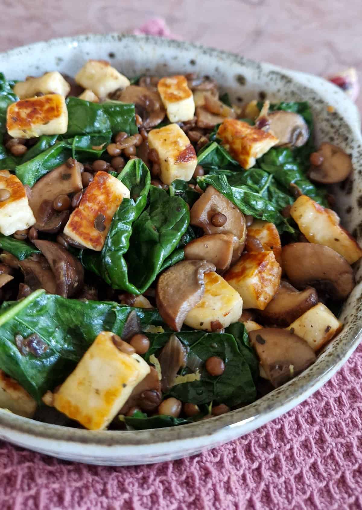Een kom gevuld met een palmkool recept met halloumi, linzen en champignons. De kom ligt op een oudroze doek.