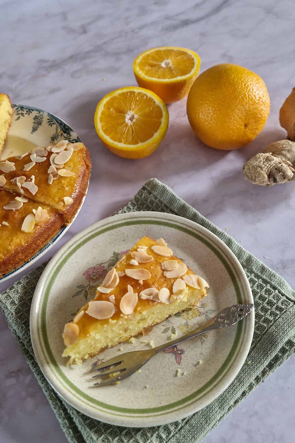 Een punt polenta cake op een taartbordje. Op de achtergrond zijn de smaakmakers van de cake te zien: sinaasappel en gember.