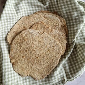Een mand gevuld met zelfgemaakte volkoren wraps. De wraps liggen op een geblokte groene theedoek.