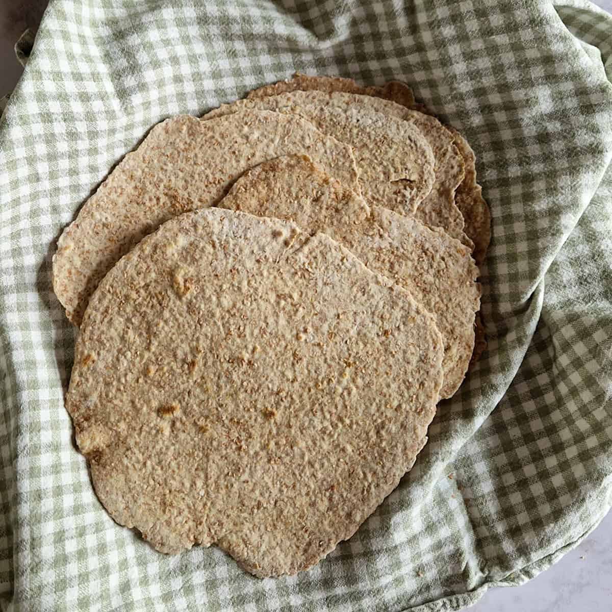 Een mand gevuld met zelfgemaakte volkoren wraps. De wraps liggen op een geblokte groene theedoek.