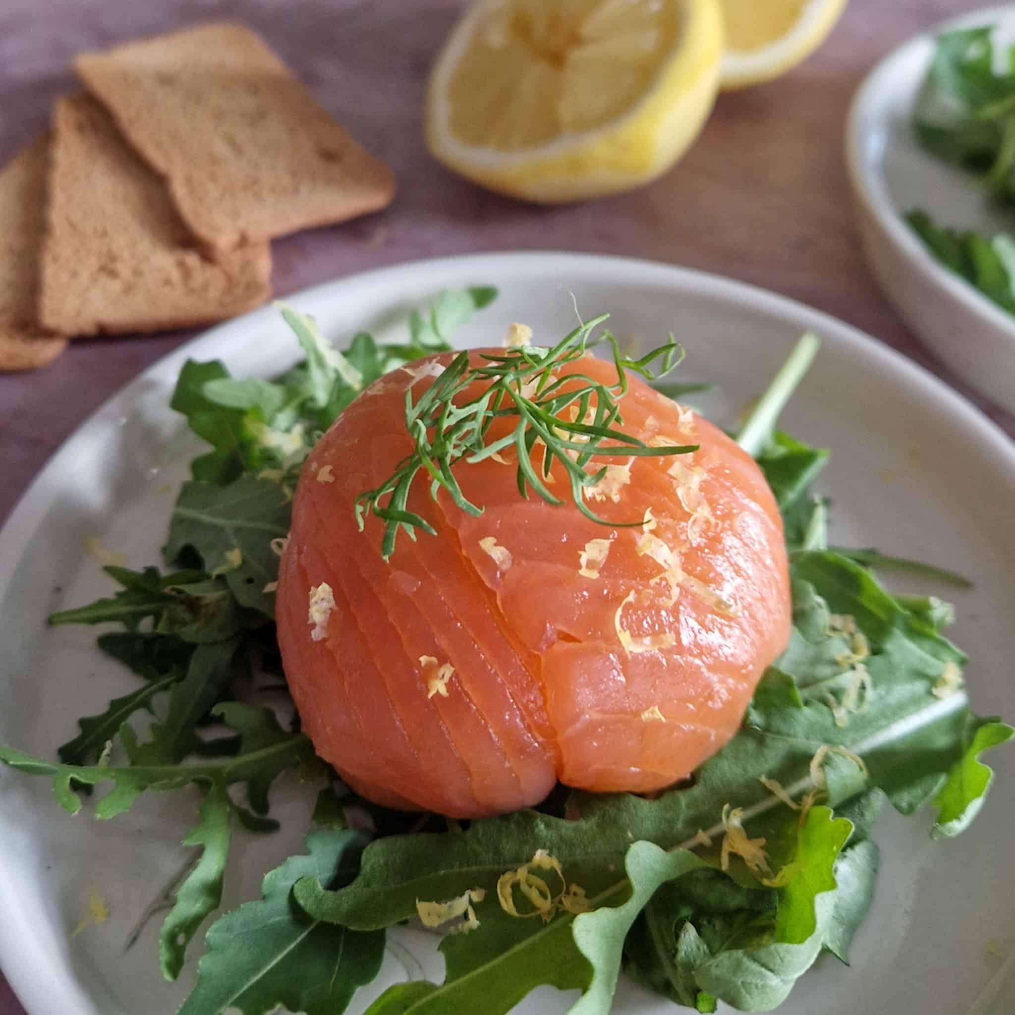 Een zalmbonbon gegarneerd met dille en citroenrasp, op een bedje van sla. Op de achtergrond liggen toastjes en een gehalveerde citroen.