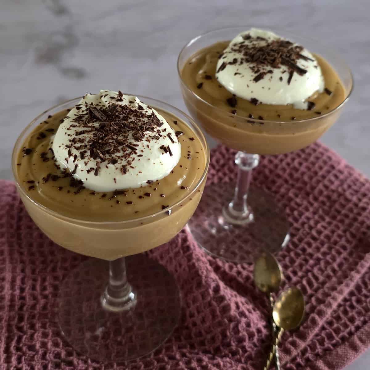 Twee glazen coupes gevuld met koffievla, slagroom en geschaafde chocolade. De glazen staan op een paars-roze doek.