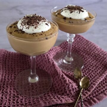 Twee glazen coupes met hopjesvla. De zelfgemaakte vla is gegarneerd met slagroom en chocoladeschaafsel.