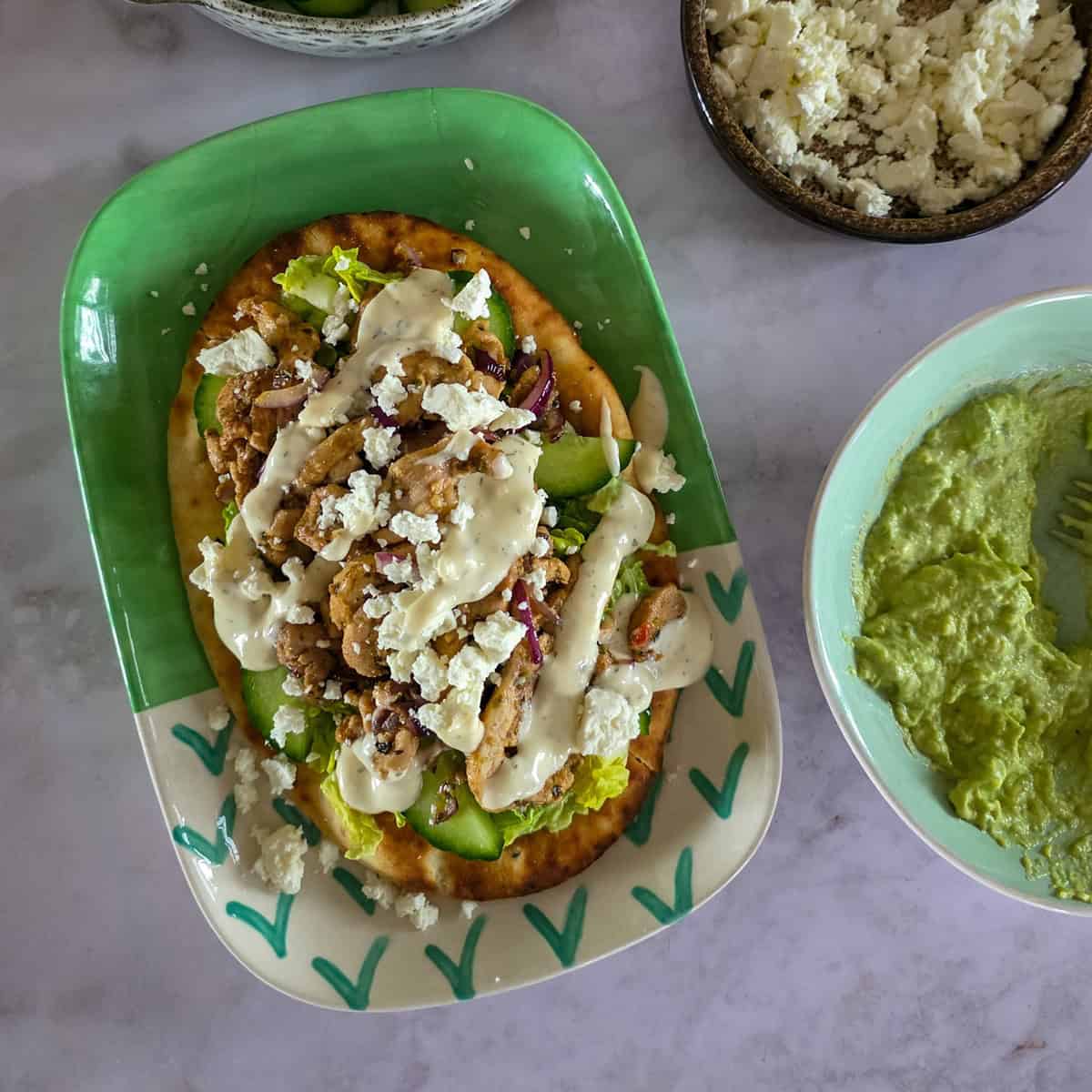 Een flatbread met kip gyros. Het broodje kip gyros ligt op een versierd groen bord en is omringd door bakjes met ingrediënten waarmee het broodje is belegd.