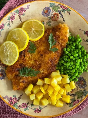 Een zelfgemaakte kipschnitzel op een ouderwets bord, met gebakken aardappeltjes en doperwten. De schnitzel is gegarneerd met citroenschijfjes en verse peterselie.